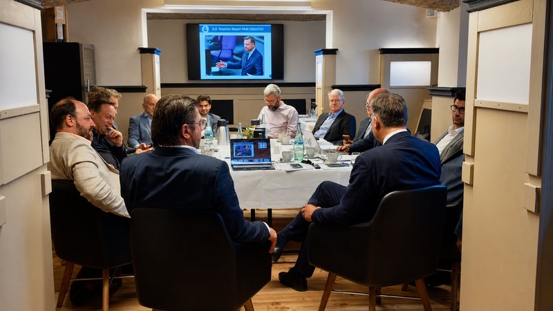 Vertreterinnen und Vertreter der Mitgliedsverbände sitzen an einem runden Konferenztisch, auf der Leinwand läuft ein Vortrag.