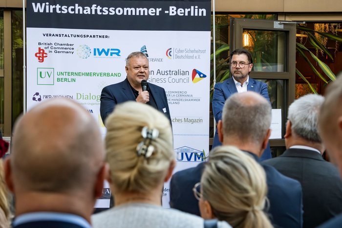 Zwei Redner sprechen vor Publikum, im Hintergrund eine Wand mit den Logos der Veranstaltungspartner.
